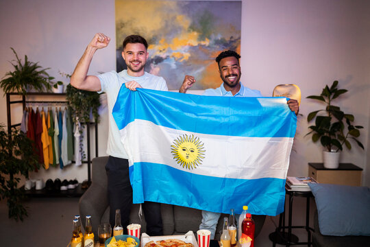 Cheerful young men gesturing fists standing with Argentinian flag at home