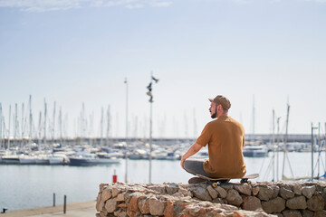 Fototapeta premium Man in cap relaxed sitting on a skateboard looking out over the harbor