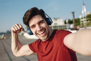 Close up young strong sporty fit sportsman man in t-shirt headphones do selfie shot mobile cell phone show muscles warm up train at sunrise sun dawn over sea beach outdoor on pier seaside in morning.