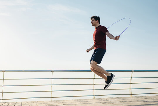 Full size young strong sporty toned fit sportsman man 20s in sports clothes jumping on skipping rope warm up training at sunrise sun dawn over sea beach outdoor on pier seaside in summer day morning