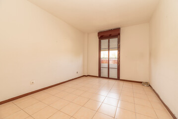 Empty room with plain white painted walls, ceramic tiled floor and reddish aluminum joinery on balcony doors