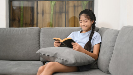 Obraz premium Smiling preteen girl relaxing on comfortable sofa and reading book