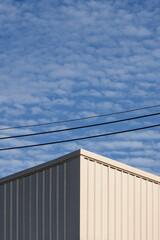 Obraz premium Sunlight on surface of white corrugated steel industrial building with power lines against fluffy clouds on blue sky background in vertical frame