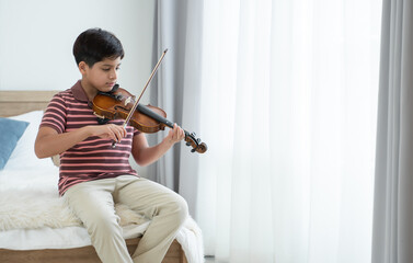 Indian handsome kid is actively practicing playing wooden violin at home. Attractive Asian teenage boy learning violin instrument alone, sitting on bed by the window in bedroom. Education concept © Pruksachat