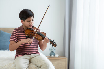 Indian handsome kid is actively practicing playing wooden violin at home. Happy Asian teenage boy learning violin instrument alone, sitting on bed by the window in bedroom. Education concept © Pruksachat