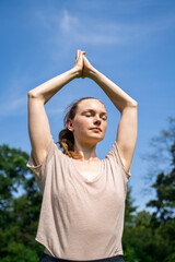 Young caucasian slim fit blond-haired woman standing in yoga pose with hands up as in prayer with closed eyes meditating, doing fitness or pilates in summer park on sunny day with clear blue sky