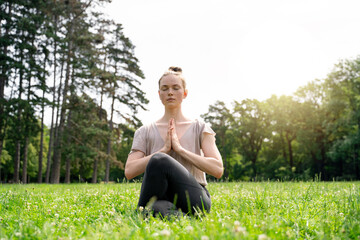 Naklejka na ściany i meble Young caucasian slim fit blond hair carefree calm woman sitting in lotus yoga pose with closed eyes and hands as in prayer, meditating on green grass meadow in summer park on early morning or sunset