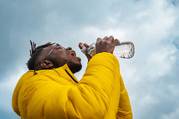 African guy with yellow windbreaker jacket drinking water from a plastic bottle against a cloudy sky