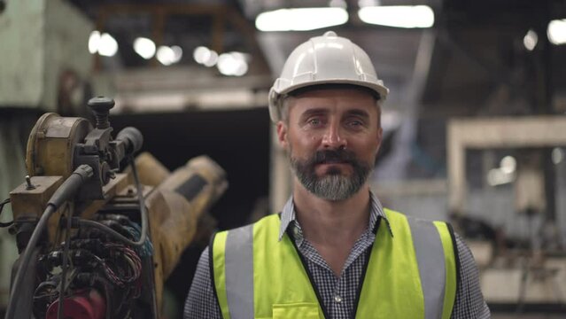 Portrait of male engineer or factory foreman worker in safety clothing holding digital tablet looking confident at camera in industry construction site with background of robotic arm