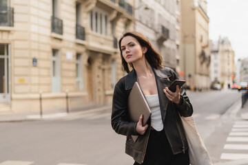 stylish freelancer in black leather jacket holding laptop and smartphone on street in paris.