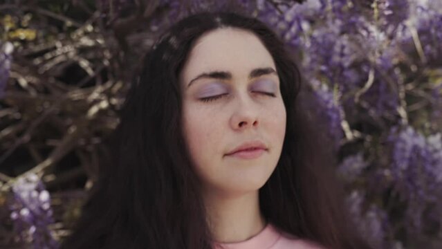 Portrait of a young beautiful Caucasian woman with her eyes closed against a background of flowering wisteria. Pan shot. Springtime.