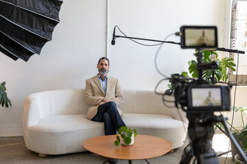 Behind the Scenes on Video Shoot of Man Being Interviewed on Camera. Caucasian Business Man Sitting on Couch with Microphone, Lighting and Camera in Frame. - obrazy, fototapety, plakaty