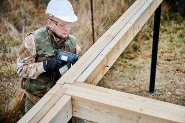 Man building wooden frame house on pile foundation. Male worker drilling hole by electric drill in timber framing of future house. Builder wearing helmet and protective goggles. Carpentry concept.