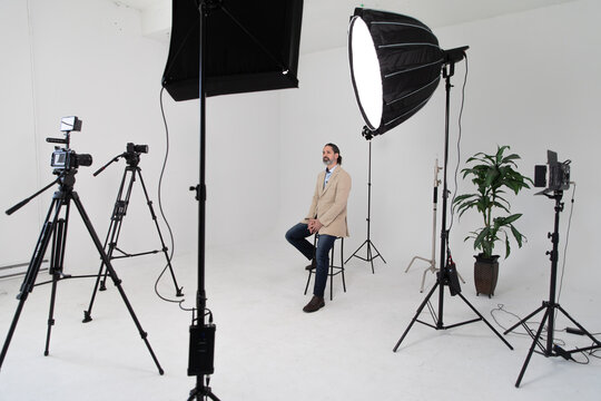 Video Production Set Behind the Scenes. Video Cameras, Lighting and Equipment Surrounds Caucasian Male Interview Subject in Middle of Frame. White Background with a Single Plant
