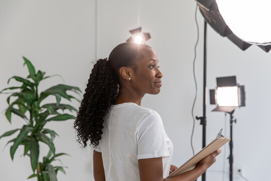 African American Woman Working On Film Set Holding Clipboard With Lighting And Equipment In Background. Proud Female Director In Video Production Studio