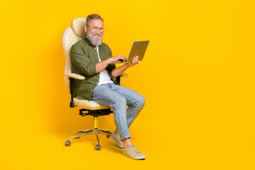 Full length portrait of cheerful aged man sitting chair hold use wireless netbook isolated on yellow color background