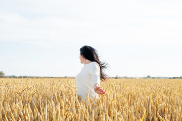 Naklejka na ściany i meble Mid adult woman in white dress standing on a wheat field with sunrise on the background, back view