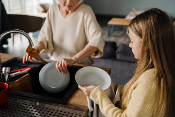 Happy grandmother and granddaughter washing dishes together in kitchen