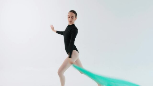 A charming ballerina in the studio performs pirouettes and jumps on one leg with a fluttering turquoise fabric in her hands
