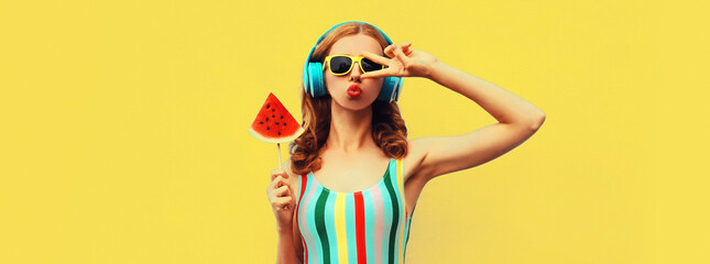 Summer portrait of stylish woman in headphones listening to music blowing her lips with juicy lollipop or ice cream shaped slice watermelon on yellow background, blank copy space for advertising text
