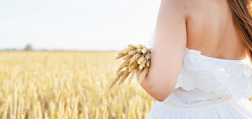 Young woman in white dress and summer hat standing on a wheat field with sunrise on the background, back view