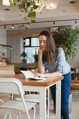 Young student brunette woman reading a book and studying at cafe with modern green interior design. Female student at coworking space or library or cafe, back to school education concept