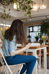 Young beautiful woman using mobile phone to browse social media sitting at cafe or coworkring space, smiling. Candid lifestyle photo of female using technology