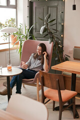 Young beautiful woman using mobile phone to browse social media sitting at cafe or coworkring space, smiling. Candid lifestyle photo of female using technology