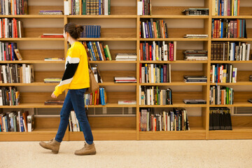 Young female student walking past wooden bookshelves in university library. Woman in public library walking inside the building. Woman in jeans and yellow sweatshirt at bookstore.