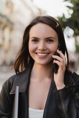 portrait of cheerful woman in black leather jacket holding laptop and talking on smartphone on street in paris.