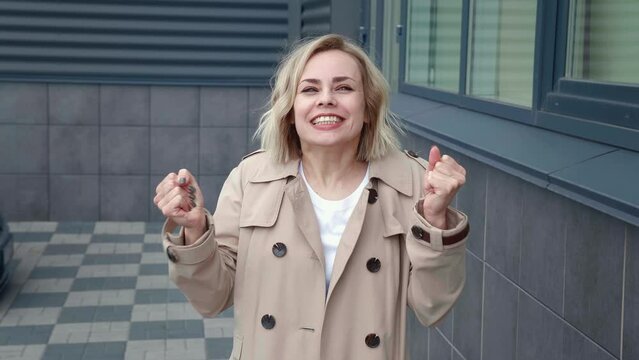 Cheerful young blonde woman in jacket making winner gesture with two hands and screaming celebrating success win, outdoors on office building background.