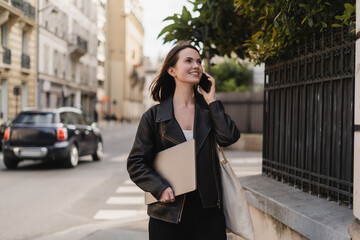 cheerful freelancer in black leather jacket holding laptop and talking on smartphone on street in...