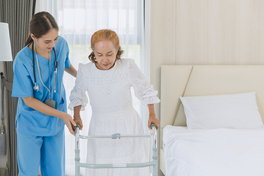 Asian elderly senior woman using walker aid with nurse care support step walking at home.