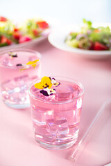 Glasses of pink violet purple cocktail decorated flowers viola pansy and fresh salad on the background.