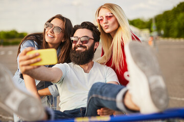 Cheerful ethnic guy sitting in pushcart and taking selfie with female friends