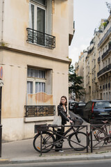 young stylish woman in black leather jacket standing near bicycle on street in paris.