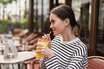 side view of smiling young woman in striped long sleeve shirt holding glass of fresh orange juice in outdoor cafe in paris.