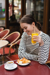 happy woman in striped long sleeve shirt holding glass of fresh orange juice and looking at croissant in outdoor cafe in paris.