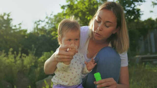 Summertime. Happy young mother hugs her little baby girl and blows soap bubbles. Slow motion. Happy Children's Day.