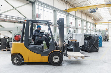 Happy mature man fork lift truck driver lifting pallet in storage warehouse and looking at camera.