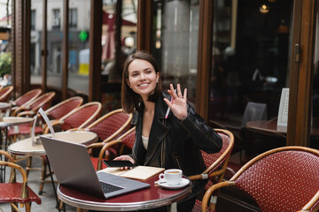 happy woman in black jacket holding smartphone and waving hand near laptop and cup of coffee in french outdoor cafe.