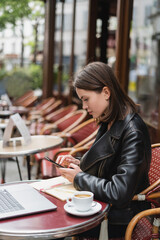 side view of freelancer in black jacket using smartphone near laptop and cup of coffee in french outdoor cafe.
