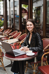 Fototapeta na wymiar happy freelancer in black jacket looking at camera near laptop and cup of coffee on table in french outdoor cafe.