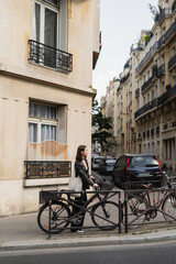 side view of young stylish woman in black leather jacket standing near bicycle on street in paris.