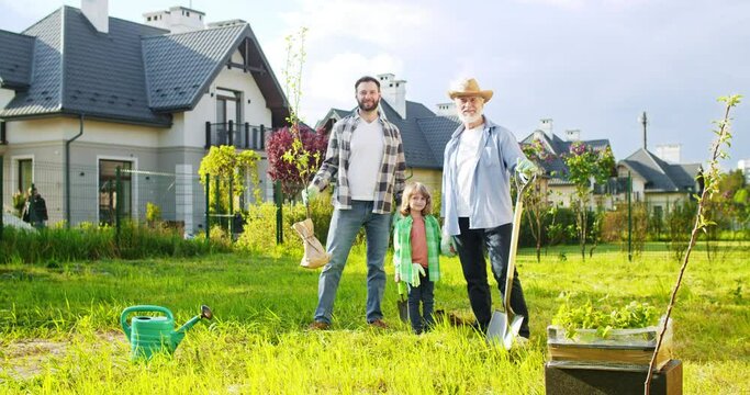 Portrait of happy family at cottage in village. Old father, son and grandson planting tree at house. Summertime day in countryside. Three generations. Outdoors.