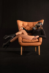 Seductive fetish model in a full gas mask, black sexy bodysuit and high stripper heels is sitting on a brown leather chair like a boss in a pretentious pose. Fetish, BDSM, bio or radiation hazard