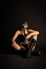 Fototapeta na wymiar Seductive girl fetish model with a slim figure poses sitting in a black gas mask, black leather or latex sexy underwear and leather boots on a black background. BDSM, perversion, fetish, air pollution