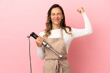 Young woman using hand blender over isolated pink background doing strong gesture