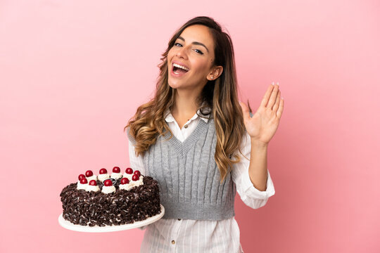 Young woman holding birthday cake over isolated pink background saluting with hand with happy expression