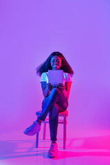 Attractive millennial black female working on tablet pc, chatting or video conferencing on web in neon light, copy space